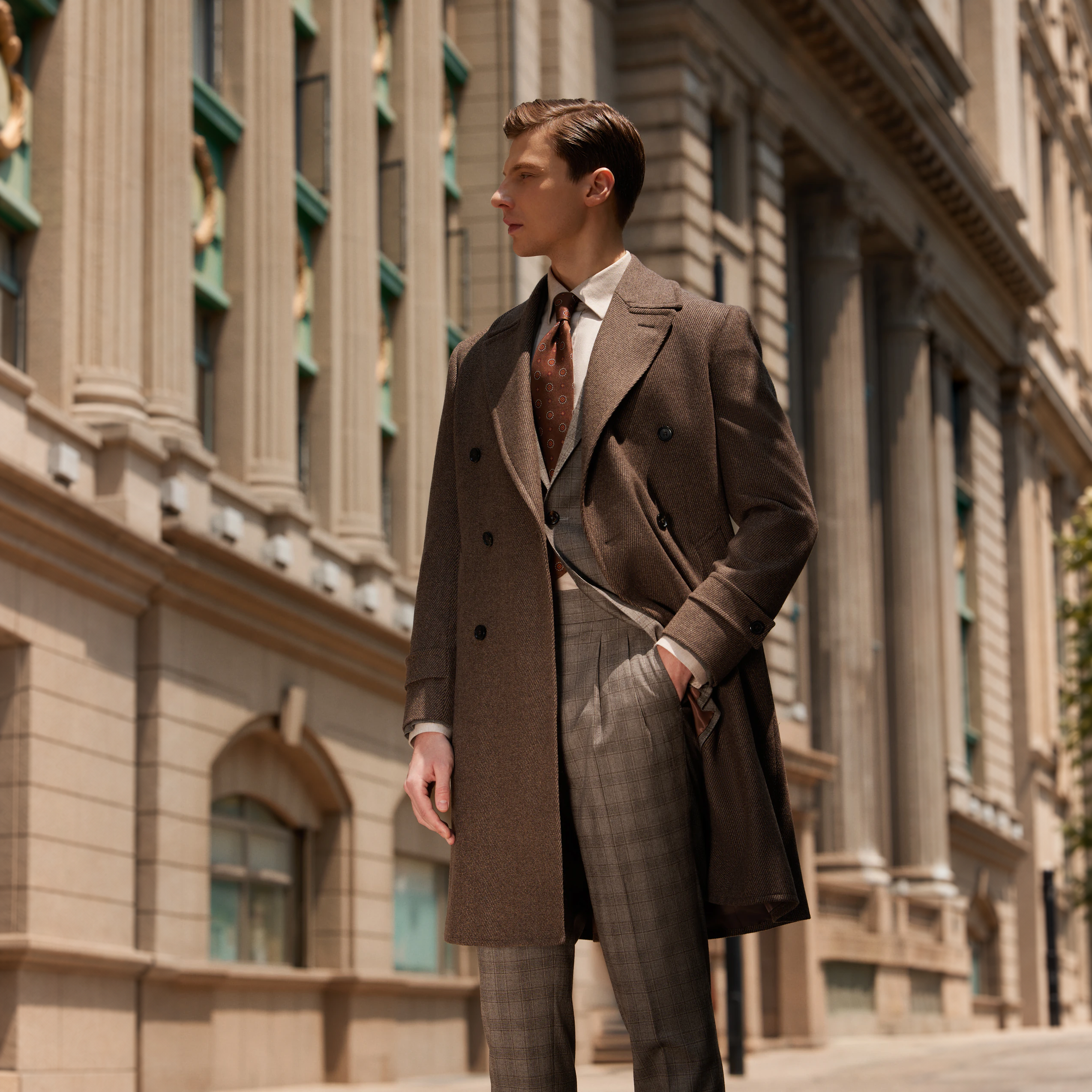 A man wearing a brown bespoke overcoat over a checked suit beside a stone building