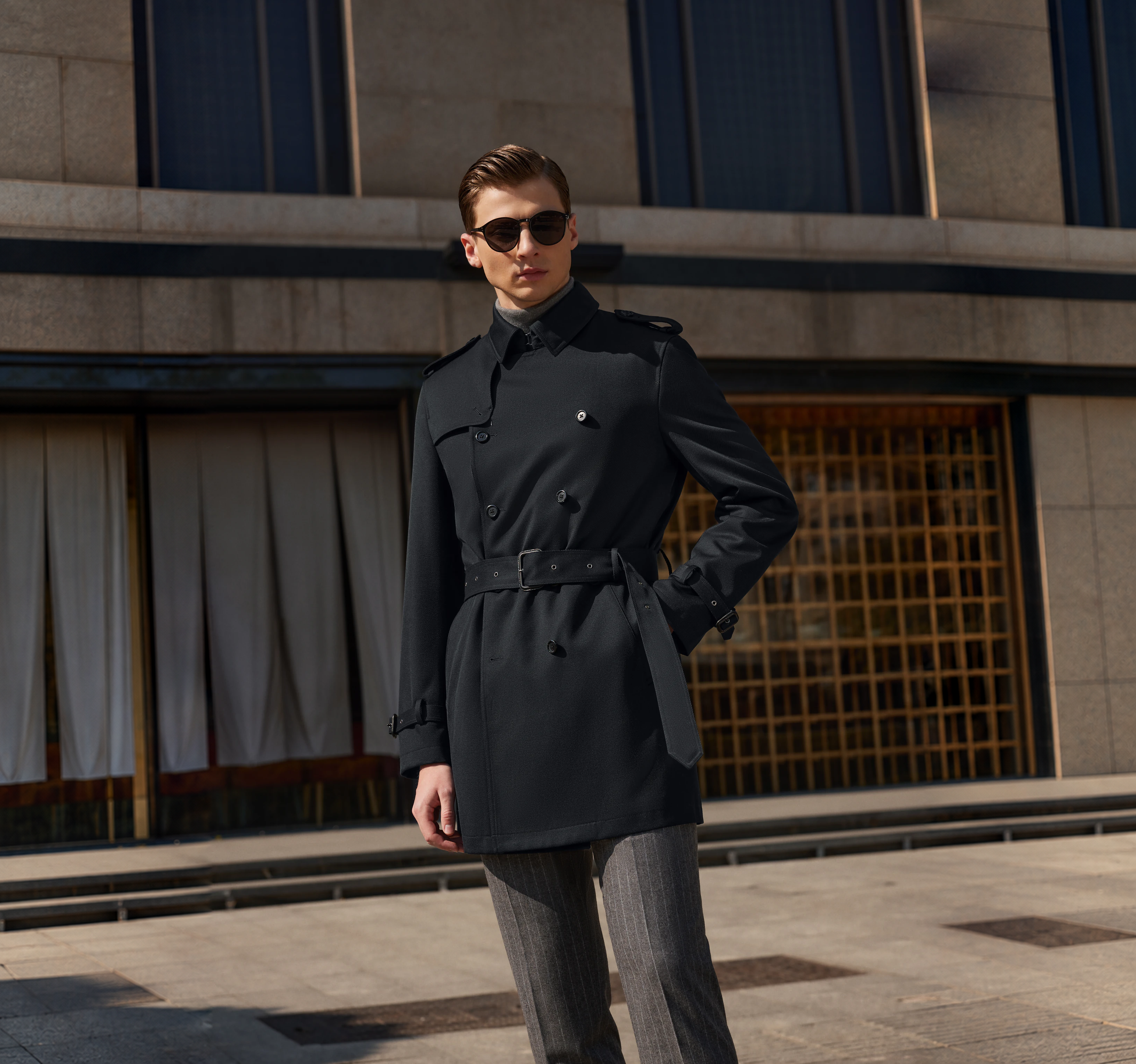 A man wearing a dark belted overcoat over a grey turtleneck and pinstripe trousers
