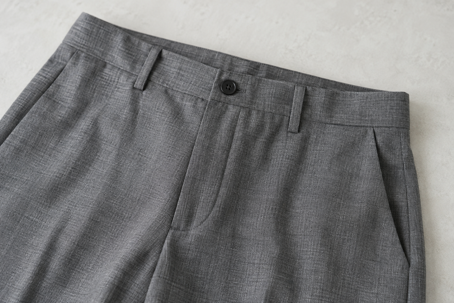 Mid-grey trouser waistband, button, fly and slanted pocket on a pale surface